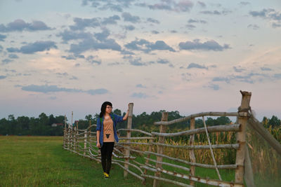 Teenage girl standing by bamboo fence on field against sky during sunset