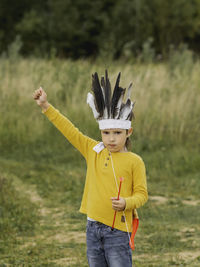 Boy plays american indian on field. kid has handmade headdress made of feathers and bow with arrows.