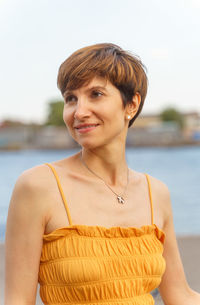 Portrait of cheerful middle-aged lady standing by river in city embankment, enjoing summer