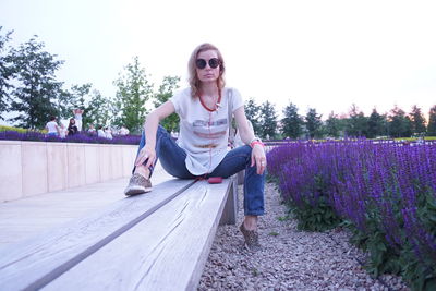 Portrait of woman sitting on bench by plants against sky