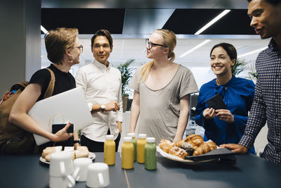 Colleagues discussing while standing by food and drink on table in office