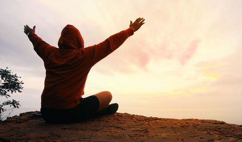 Rear view of hiker with arms raised sitting on cliff against sky during sunset