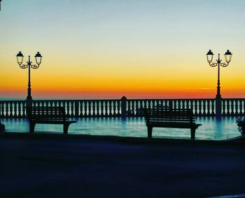 Bench on street by sea against sky during sunset