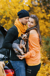 A guy and a girl in love embrace in an autumn park, a guy holds a cute dog in his arms