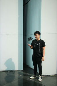 Full length of young man catching camera while standing against wall