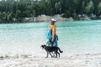 Rear view of young woman traveler walking on sandy shore of blue lake or river with fluffy gray dog