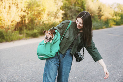 A smiling girl with a camera around her neck holds a green backpack on her shoulder.