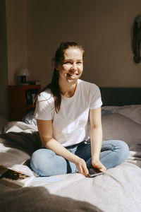 Happy young woman with smart phone sitting on bed at home