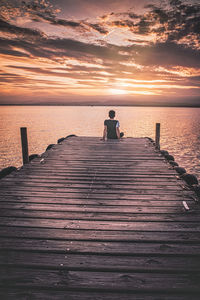 Rear view of man sitting on pier over sea against sky during sunset