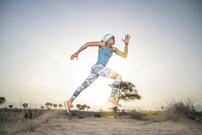 Full length of woman jumping while running in desert against clear sky during sunset