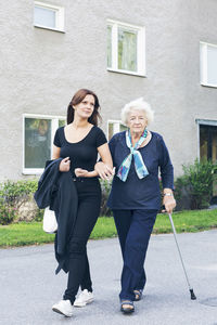 Portrait of elderly woman holding cane while walking with granddaughter on footpath against building