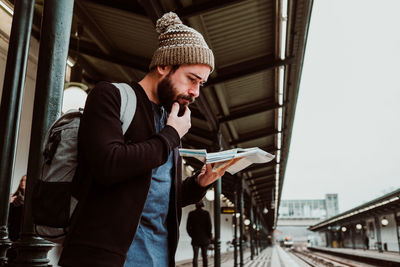 Side view of man wearing knit hat holding map standing at railroad station platform