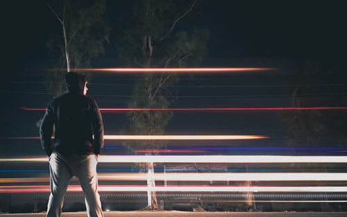 Rear view of man standing in front of moving vehicles making light trails