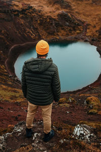 Back view of young tourist in winter wear looking at pound and mountain river between stone hill