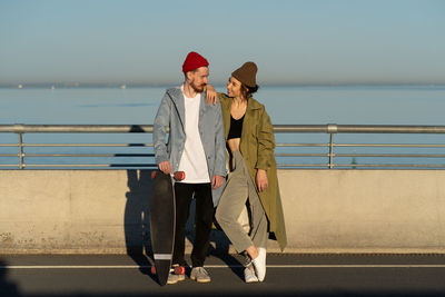 Happy couple of lovers hipsters with longboard on bridge in sunshine smiling wearing stylish clothes