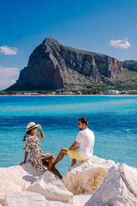 Woman sitting on rock by sea against mountains