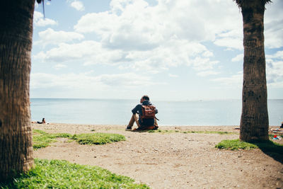 Rear view of man sitting at beach against cloudy sky during summer