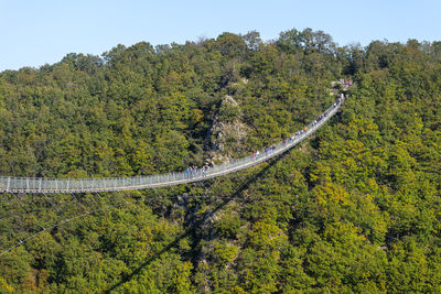 A hanging wooden bridge with steel ropes seen from the side against a blue sky, visible tourists. 