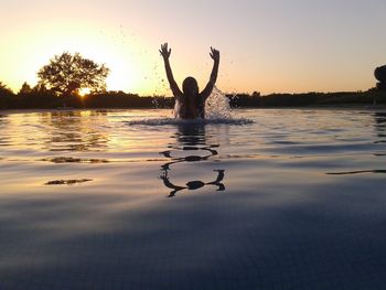 Playful woman splashing water in swimming pool against sky during sunset