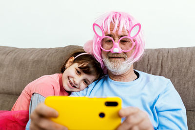 Happy child sitting on couch with grandfather in big pink plastic glasses and pink wig with false ears and making selfie on smartphone in room