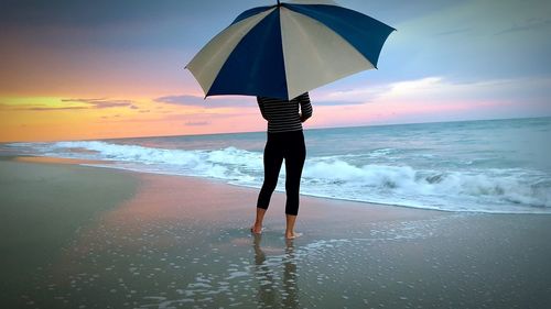 Woman with umbrella standing at beach against sky during sunset