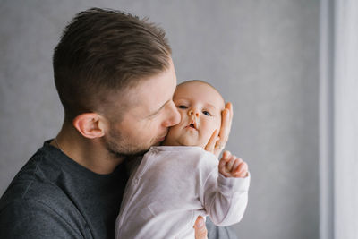 Dad holds his baby in his arms at home and kisses the baby on the cheek