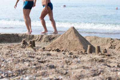Sand pyramid on the beach by the sea, two people walking along the edge of the water in the