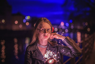 Portrait of young woman holding illuminated string lights by lake at night