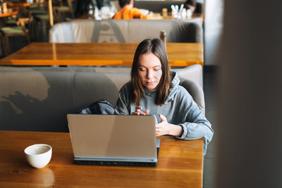 Adult charming brunette woman in casual clothes working with laptop using mobile phone at cafe