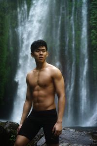 Portrait of shirtless man standing against waterfall