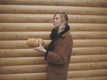 Side view of woman holding pumpkin while standing by wooden wall