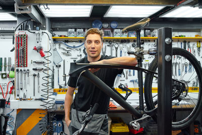 Happy male mechanic smiling and looking at camera while leaning on bike under repair against wall with tools in garage