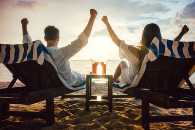 Rear view of couple with arms raised relaxing on lounge chairs at beach