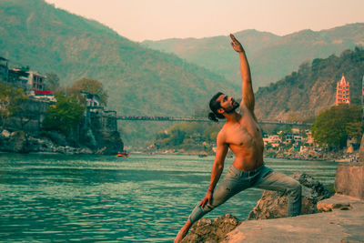 Shirtless man practicing yoga by river against mountains