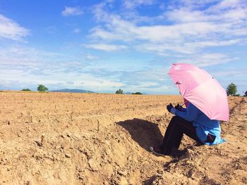 Side view of woman with umbrella sitting on field against sky during sunny day