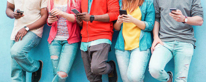 Midsection of friends using smart phones while standing against wall