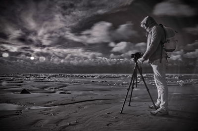 Side view of man with camera and tripod photographing at beach against cloudy sky