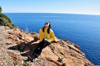 Young woman sitting on rock by sea against sky