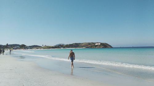 Rear view of shirtless man walking at beach against clear blue sky