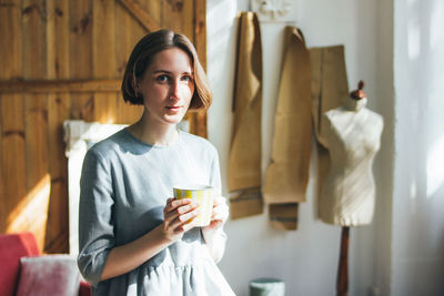 Portrait of young woman drinking coffee while standing against wall