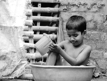 Shirtless boy taking shower in bucket against wall