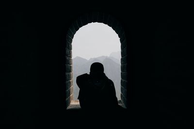 Rear view of silhouette man at arch window against mountains and sky