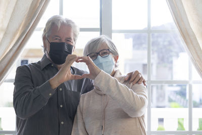 Portrait of senior couple wearing masks making heart shape against window