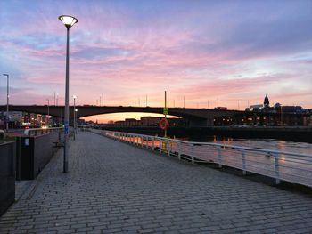 Illuminated street by river against sky during sunset