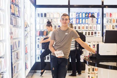 Portrait of confident young salesman holding phone cover while standing by checkout with customers in background