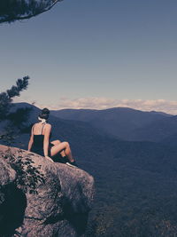 Rear view of teenage girl sitting on rock against sky