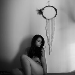 Naked young woman sitting against wall at home