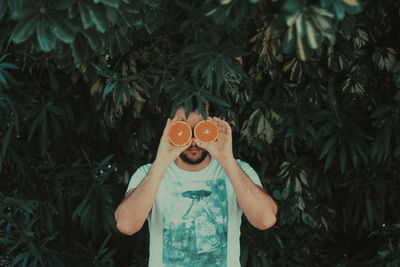 Man holding oranges in front of eyes standing against plants