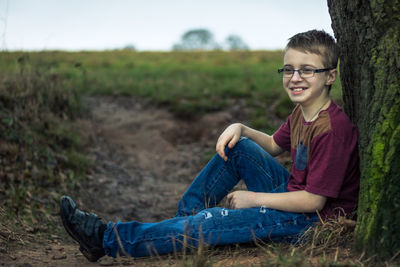 Boy smiling while sitting on field by tree