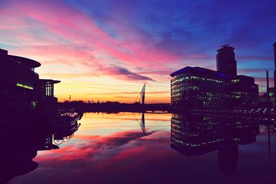 Modern buildings by lake against sky during sunset in city